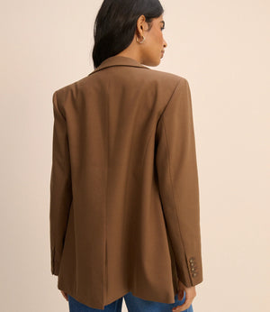A woman shown from the back wearing an oversized brown blazer with blue jeans, photographed against a plain beige studio background.