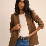 A woman modeling a brown blazer over a white top and blue jeans with a brown belt, photographed against a plain beige studio background.