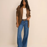 A woman modeling a brown blazer, white top, and blue wide-leg jeans with a gold-buckled belt, standing against a plain beige studio background.