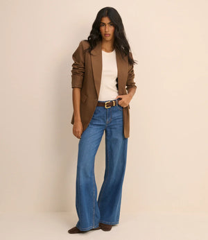 A woman modeling a brown blazer, white top, and blue wide-leg jeans with a gold-buckled belt, standing against a plain beige studio background.