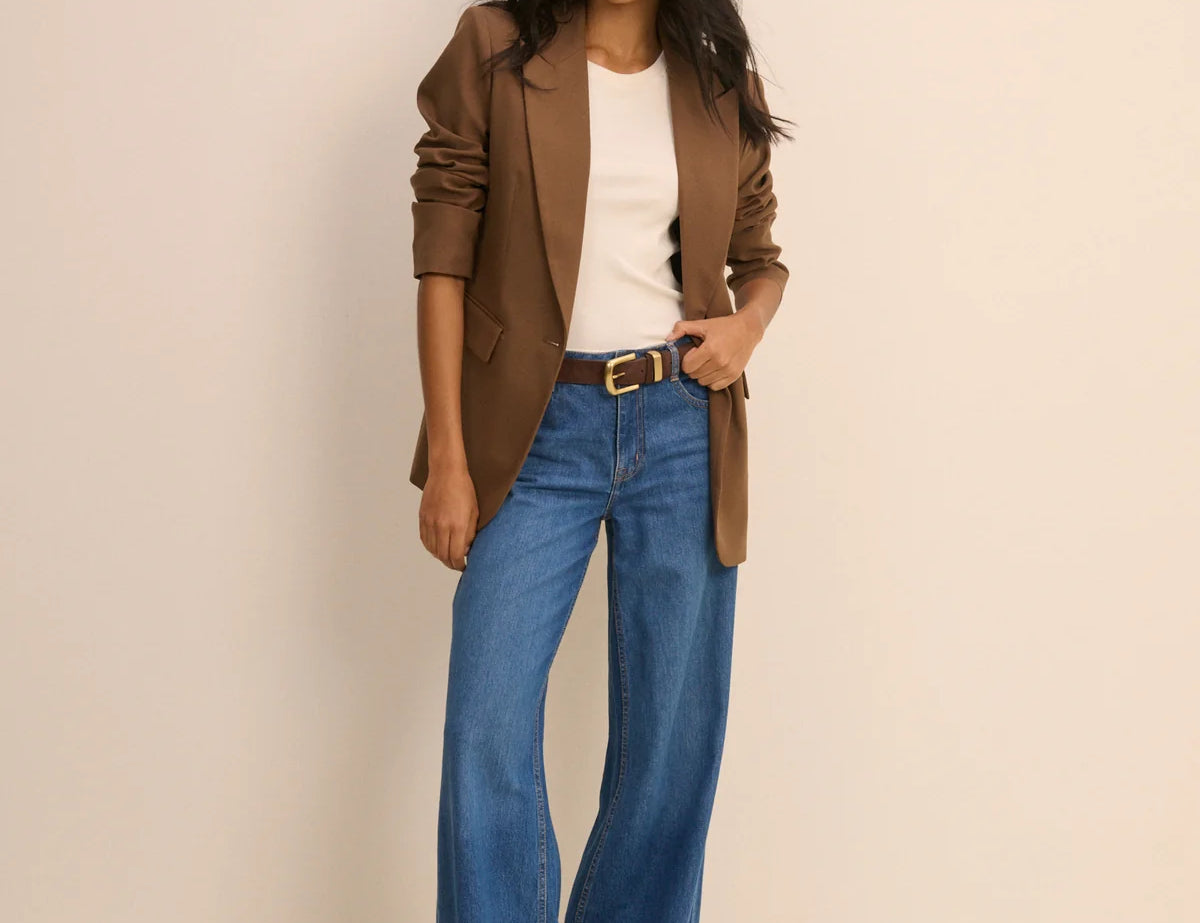 A woman modeling a brown blazer, white top, and blue wide-leg jeans with a gold-buckled belt, standing against a plain beige studio background.