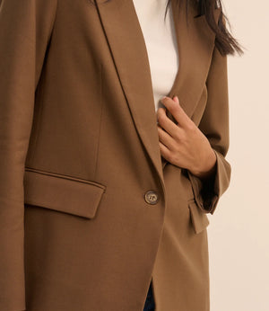 A close-up of a person wearing a camel brown blazer over a white top, with the hand adjusting the front button.