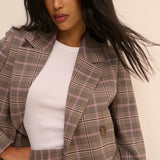 A woman wearing a brown and pink plaid blazer over a white ribbed top, photographed against a plain light beige background.