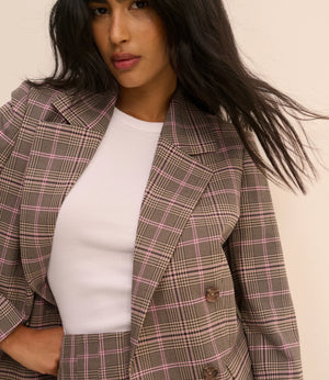 A woman wearing a brown and pink plaid blazer over a white ribbed top, photographed against a plain light beige background.
