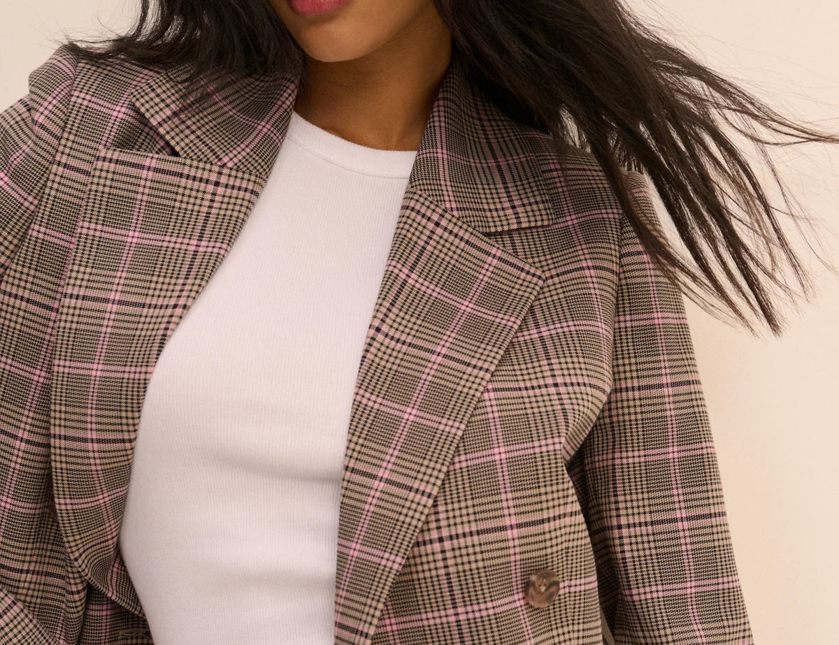 A woman wearing a brown and pink plaid blazer over a white ribbed top, photographed against a plain light beige background.