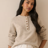 A woman wearing a light beige knit sweater with a buttoned henley neckline and white pants, posed against a plain beige wall.