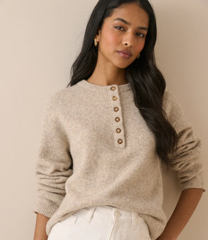 A woman wearing a light beige knit sweater with a buttoned henley neckline and white pants, posed against a plain beige wall.