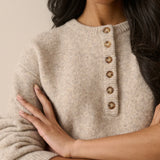 A close-up of a woman wearing a soft knit sweater in a warm light beige color, featuring a buttoned henley-style neckline with round metallic buttons.
