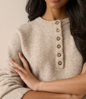 A close-up of a woman wearing a soft knit sweater in a warm light beige color, featuring a buttoned henley-style neckline with round metallic buttons.