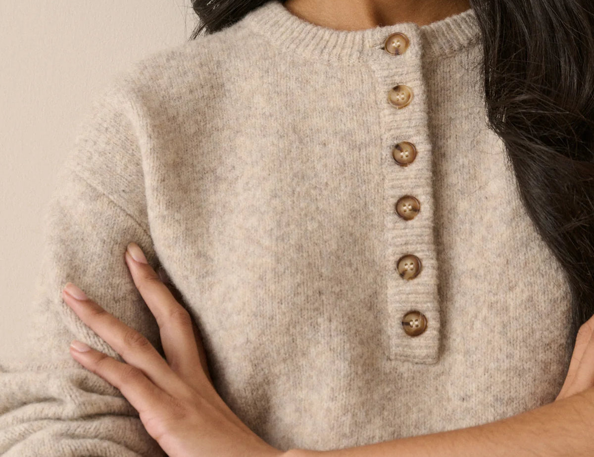 A close-up of a woman wearing a soft knit sweater in a warm light beige color, featuring a buttoned henley-style neckline with round metallic buttons.