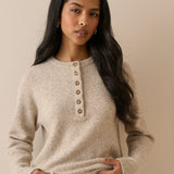 A woman modeling a light beige knit sweater with a buttoned henley neckline, paired with white pants, against a solid beige background.