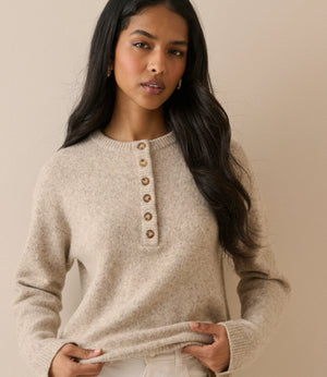 A woman modeling a light beige knit sweater with a buttoned henley neckline, paired with white pants, against a solid beige background.