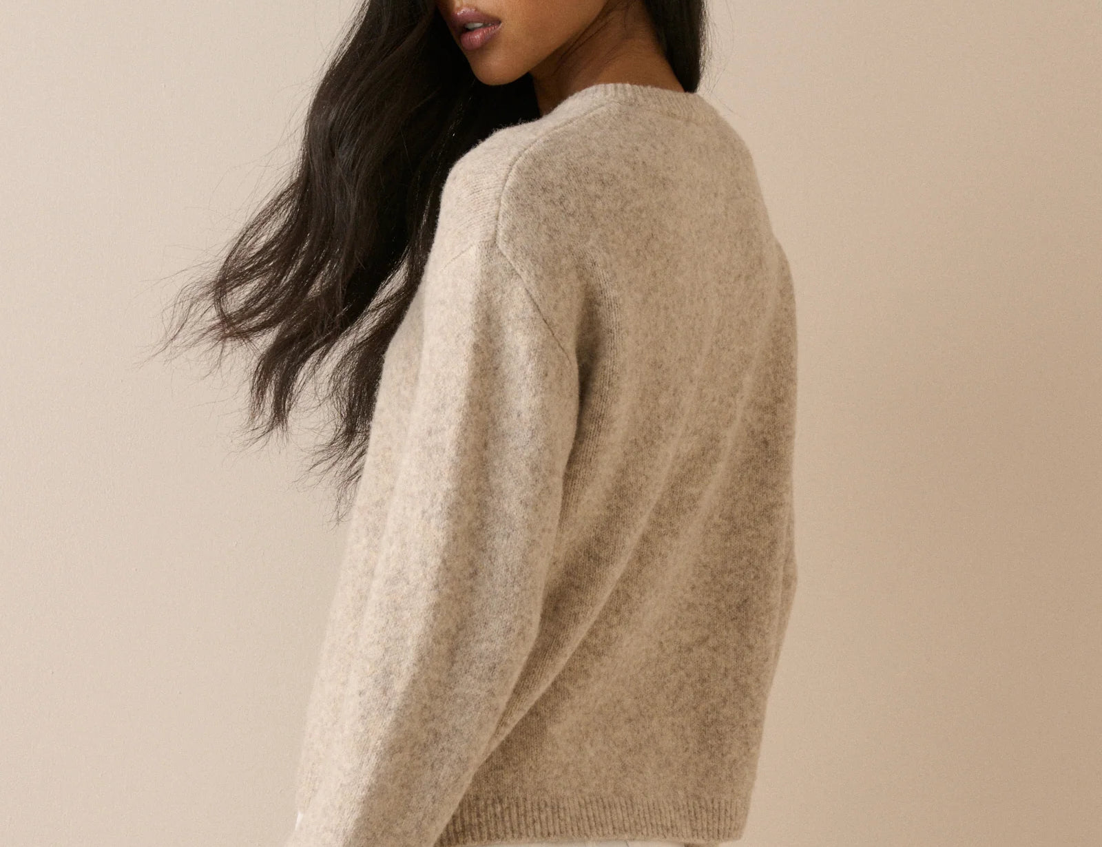 A woman with long dark hair is shown from the back wearing a fuzzy, light beige knit sweater and white pants against a solid beige background.