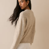 A woman with long dark hair is shown from the back wearing a fuzzy, light beige knit sweater and white pants against a solid beige background.