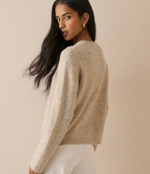 A woman with long dark hair is shown from the back wearing a fuzzy, light beige knit sweater and white pants against a solid beige background.