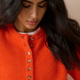A close-up of a woman wearing a bright orange knit sweater with a buttoned henley neckline over a white undershirt, set against a soft beige background.