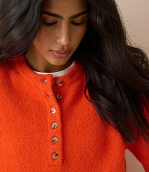 A close-up of a woman wearing a bright orange knit sweater with a buttoned henley neckline over a white undershirt, set against a soft beige background.