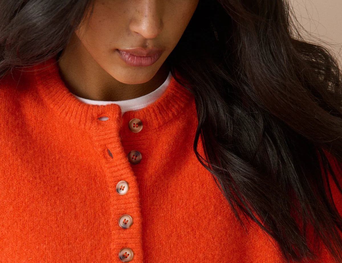 A close-up of a woman wearing a bright orange knit sweater with a buttoned henley neckline over a white undershirt, set against a soft beige background.