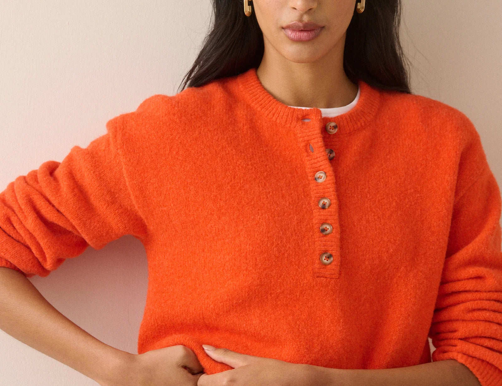 A woman wearing a bright orange fuzzy knit sweater with a buttoned henley neckline, paired with dark pleated trousers, posed against a solid light beige background.