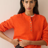 A woman wearing a bright orange fuzzy knit sweater with a buttoned henley neckline, paired with dark pleated trousers, posed against a solid light beige background.