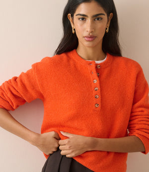 A woman wearing a bright orange fuzzy knit sweater with a buttoned henley neckline, paired with dark pleated trousers, posed against a solid light beige background.