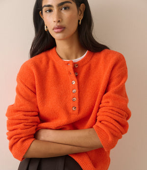 A woman wearing a bright orange fuzzy knit sweater with a buttoned henley-style neckline, photographed against a plain beige studio background.