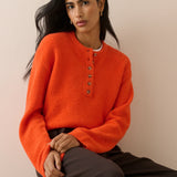 A woman wearing a bright orange fuzzy knit henley sweater with brown buttons, paired with dark brown trousers, seated against a plain beige background.
