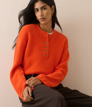 A woman wearing a bright orange fuzzy knit henley sweater with brown buttons, paired with dark brown trousers, seated against a plain beige background.