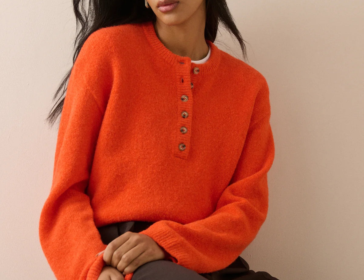 A woman wearing a bright orange fuzzy knit henley sweater with brown buttons, paired with dark brown trousers, seated against a plain beige background.
