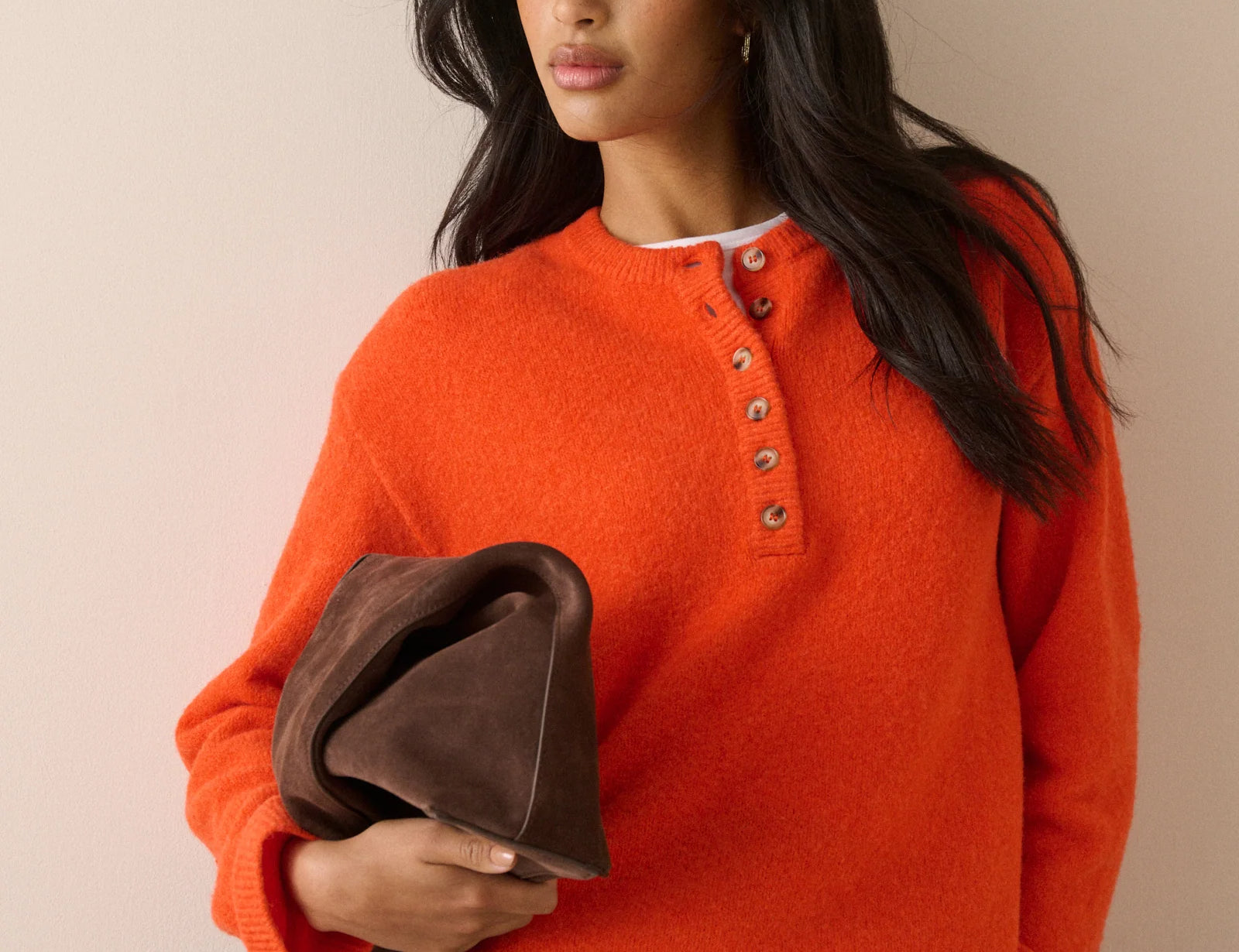 A woman wearing a bright orange knit henley sweater with brown buttons, holding a dark brown leather clutch bag against a plain beige background.