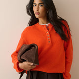 A woman wearing a bright orange knit henley sweater with brown buttons, holding a dark brown leather clutch bag against a plain beige background.