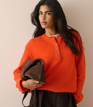 A woman wearing a bright orange knit henley sweater with brown buttons, holding a dark brown leather clutch bag against a plain beige background.