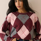 A woman wearing a cropped knit sweater with a large argyle pattern in burgundy, light pink, and gray, paired with blue jeans against a plain beige background.