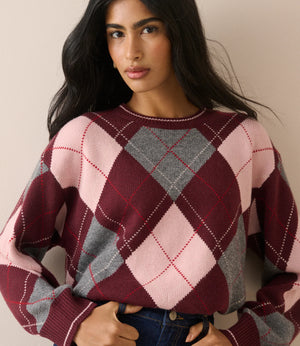 A woman wearing a cropped knit sweater with a large argyle pattern in burgundy, light pink, and gray, paired with blue jeans against a plain beige background.