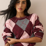 A woman wearing a pink, burgundy, and gray plaid knit sweater with rolled-up sleeves, paired with blue jeans, set against a plain beige background.