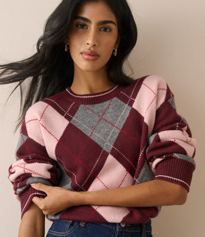A woman wearing a pink, burgundy, and gray plaid knit sweater with rolled-up sleeves, paired with blue jeans, set against a plain beige background.