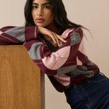 A woman wearing a pink, burgundy, and gray argyle sweater leans against a wooden table, wearing dark blue jeans against a beige background.