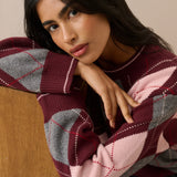 A woman wearing a chunky knit sweater with an argyle pattern in burgundy, light pink, and gray, leaning against a wooden surface.