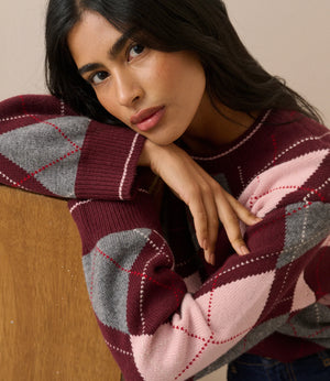A woman wearing a chunky knit sweater with an argyle pattern in burgundy, light pink, and gray, leaning against a wooden surface.