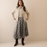 A woman stands against a plain beige studio background wearing a cream zip-front jacket over a cream lace-up blouse, a blue and gray plaid midi skirt, and dark brown ankle boots.