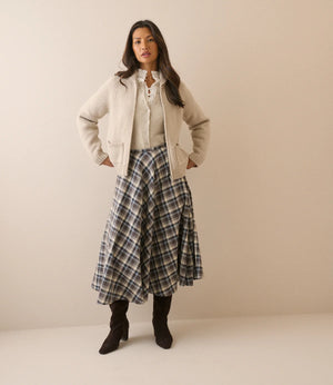 A woman stands against a plain beige studio background wearing a cream zip-front jacket over a cream lace-up blouse, a blue and gray plaid midi skirt, and dark brown ankle boots.