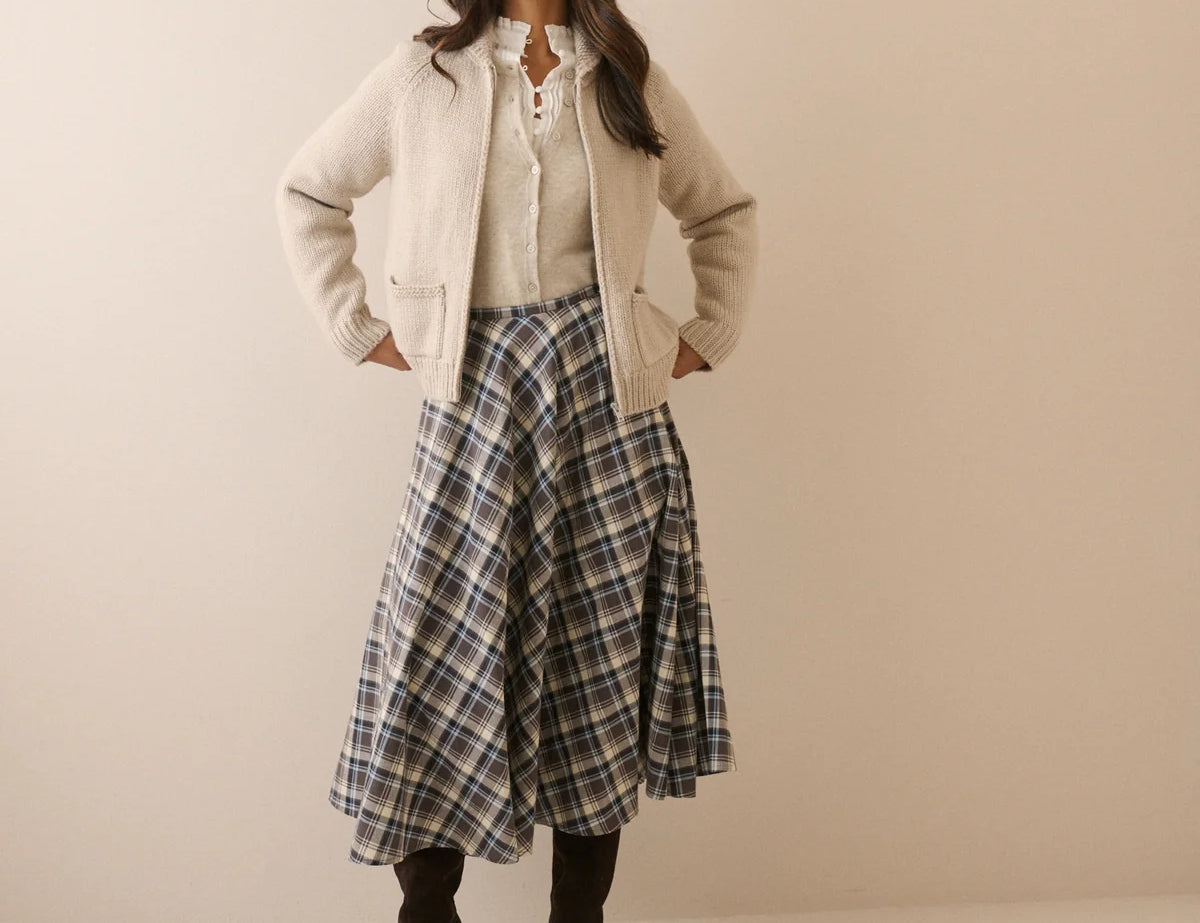 A woman stands against a plain beige studio background wearing a cream zip-front jacket over a cream lace-up blouse, a blue and gray plaid midi skirt, and dark brown ankle boots.