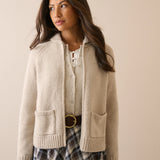 A woman wearing a cream-colored knit zip-up cardigan with front pockets over a white top and a plaid skirt, standing against a solid beige background.