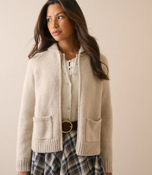 A woman wearing a cream-colored knit zip-up cardigan with front pockets over a white top and a plaid skirt, standing against a solid beige background.