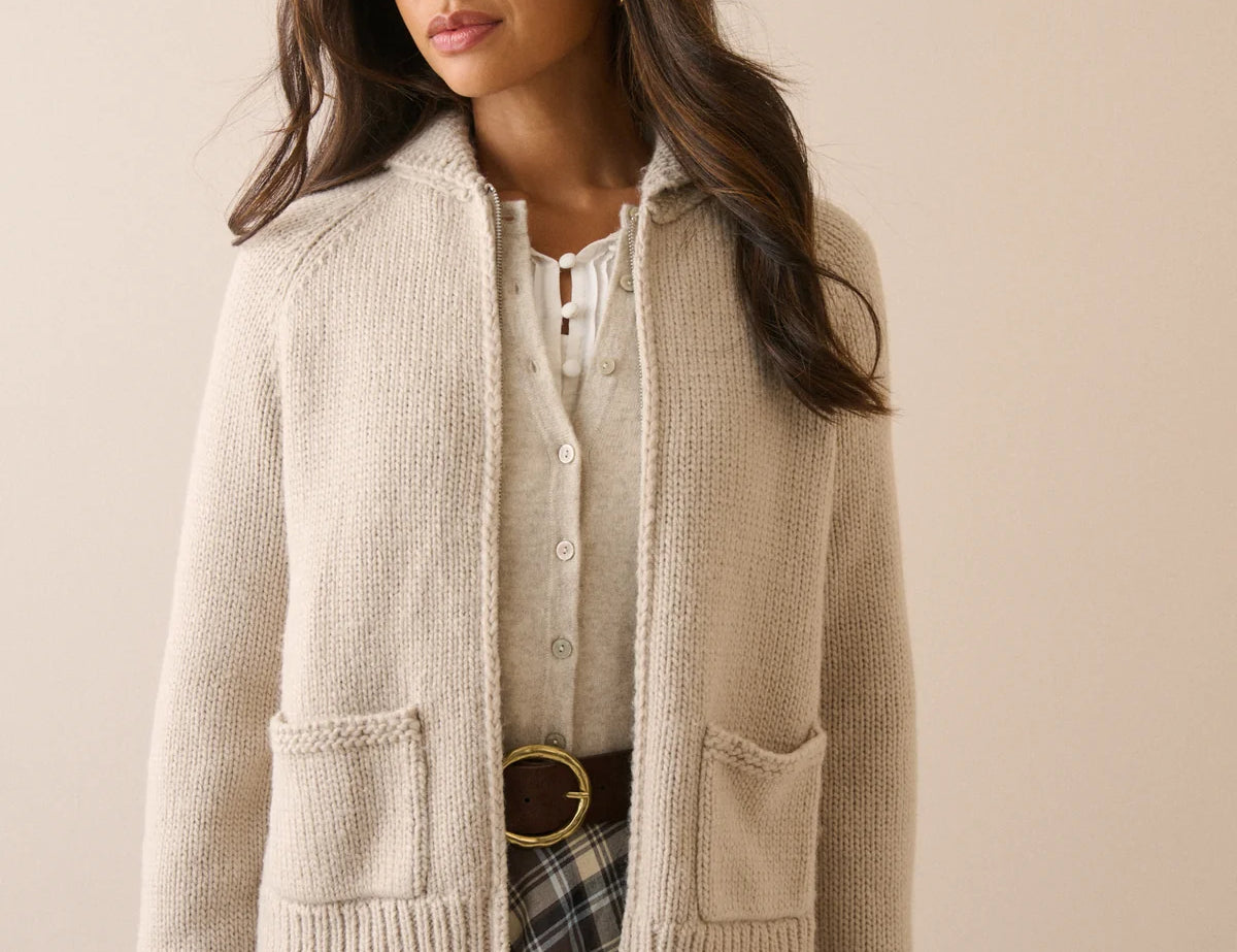 A woman wearing a cream-colored knit zip-up cardigan with front pockets over a white top and a plaid skirt, standing against a solid beige background.