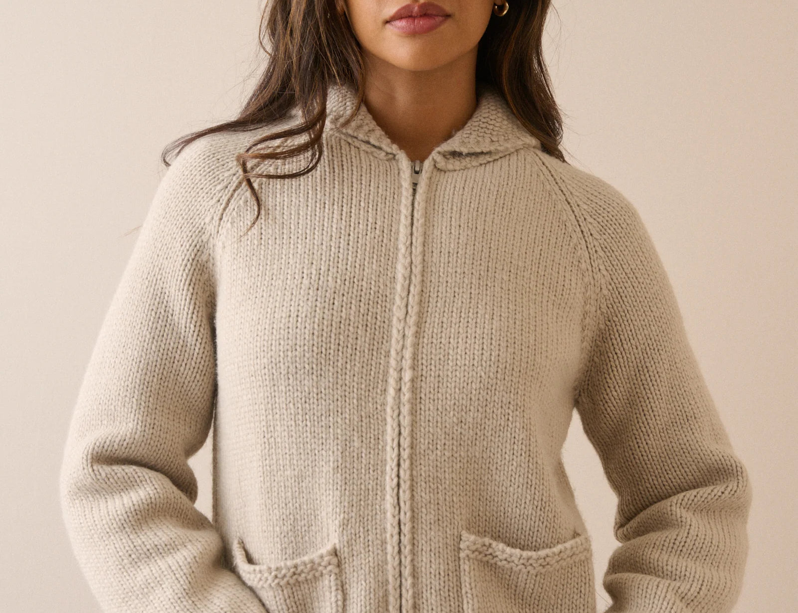 A woman wearing a beige knit zip-up hoodie cardigan with a hood and front pockets, photographed against a plain beige studio background.