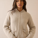 A woman wearing a beige knit zip-up hoodie cardigan with a hood and front pockets, photographed against a plain beige studio background.