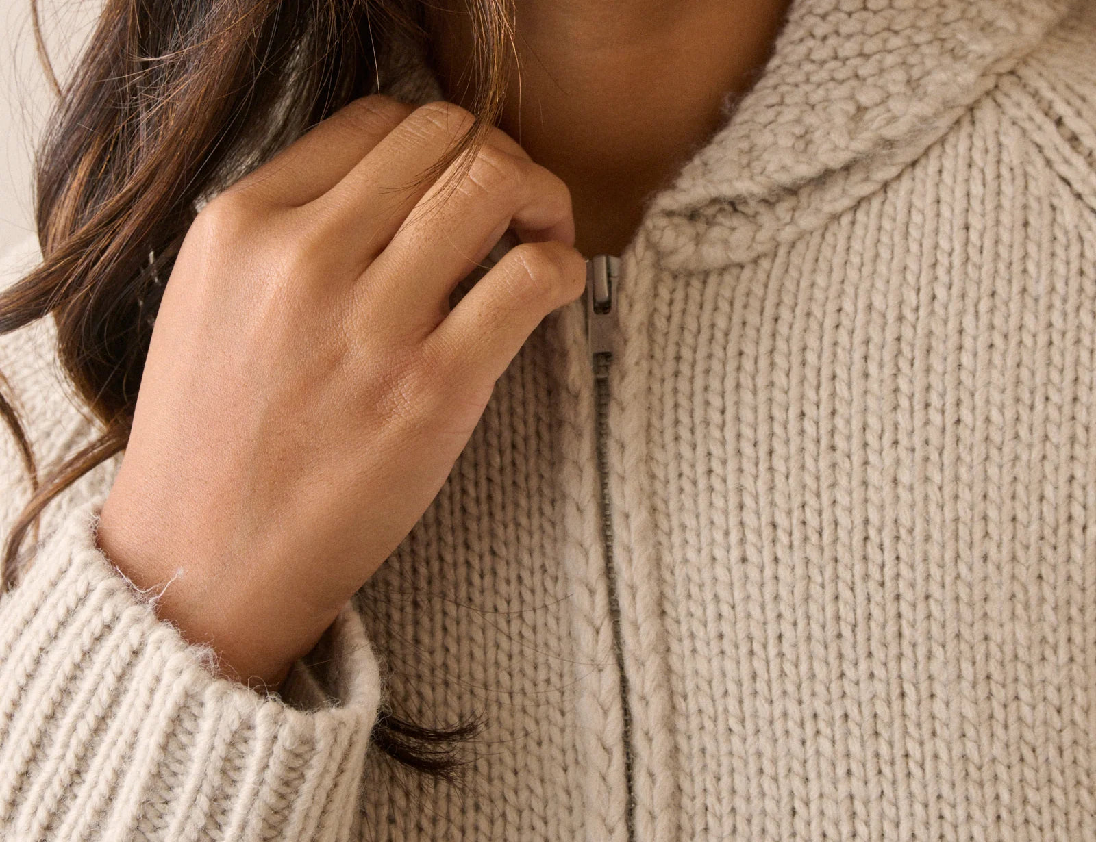 Close-up of a person wearing a chunky knit zip-up sweater in a warm off-white or cream tone, with the hand adjusting the zipper.
