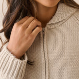 Close-up of a person wearing a chunky knit zip-up sweater in a warm off-white or cream tone, with the hand adjusting the zipper.