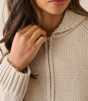 Close-up of a person wearing a chunky knit zip-up sweater in a warm off-white or cream tone, with the hand adjusting the zipper.